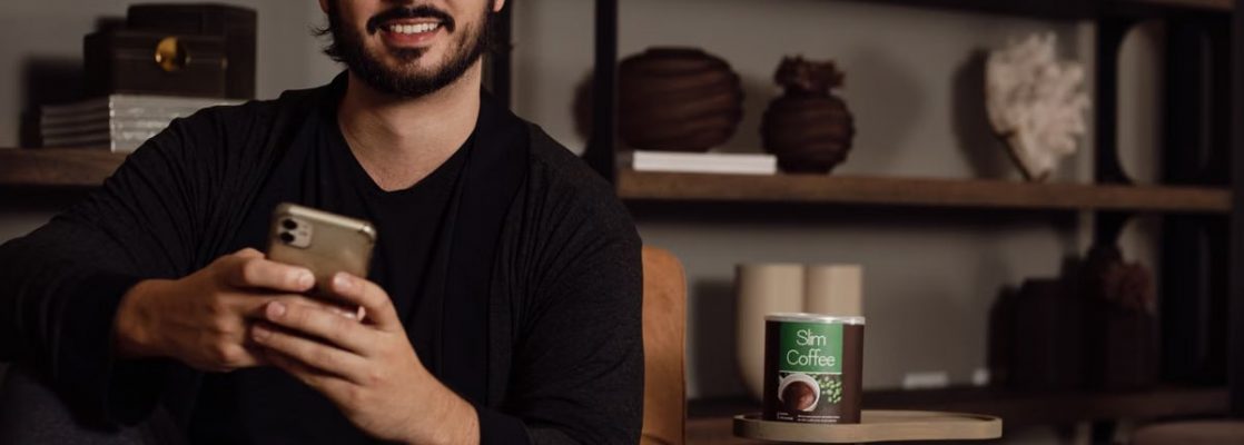 A bearded man smiling while using a smartphone, sitting on a sofa in a cozy living room setting.