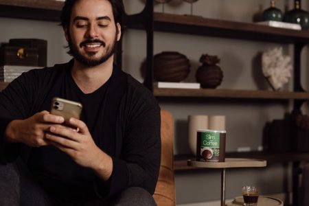 A bearded man smiling while using a smartphone, sitting on a sofa in a cozy living room setting.
