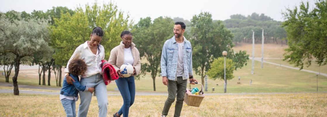 A joyful family walking through a park on a sunny day, carrying a picnic basket and a ball.