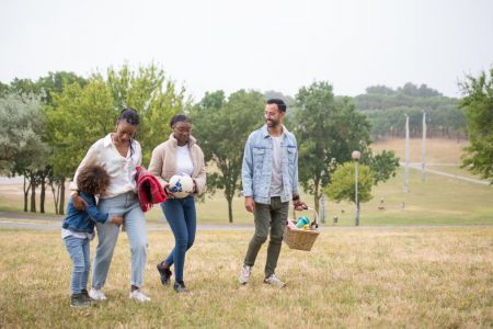 A joyful family walking through a park on a sunny day, carrying a picnic basket and a ball.